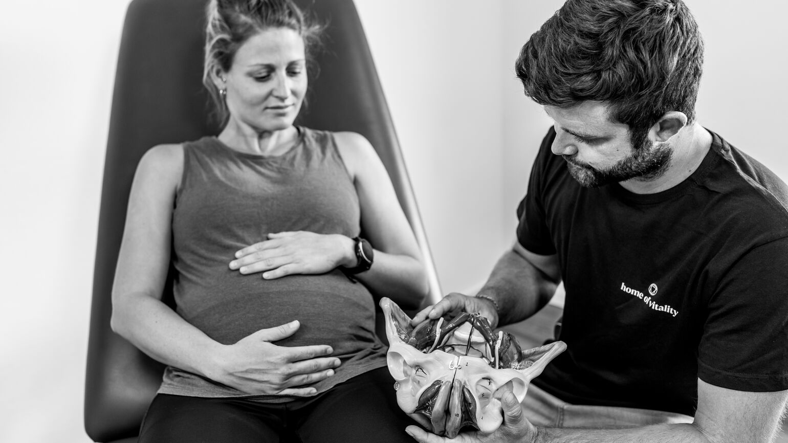 Eine Schwangere Patientin sitzt auf einer Behandlungsliege, während der Therapeut ihr ein anatomisches Beckenmodell zeigt.