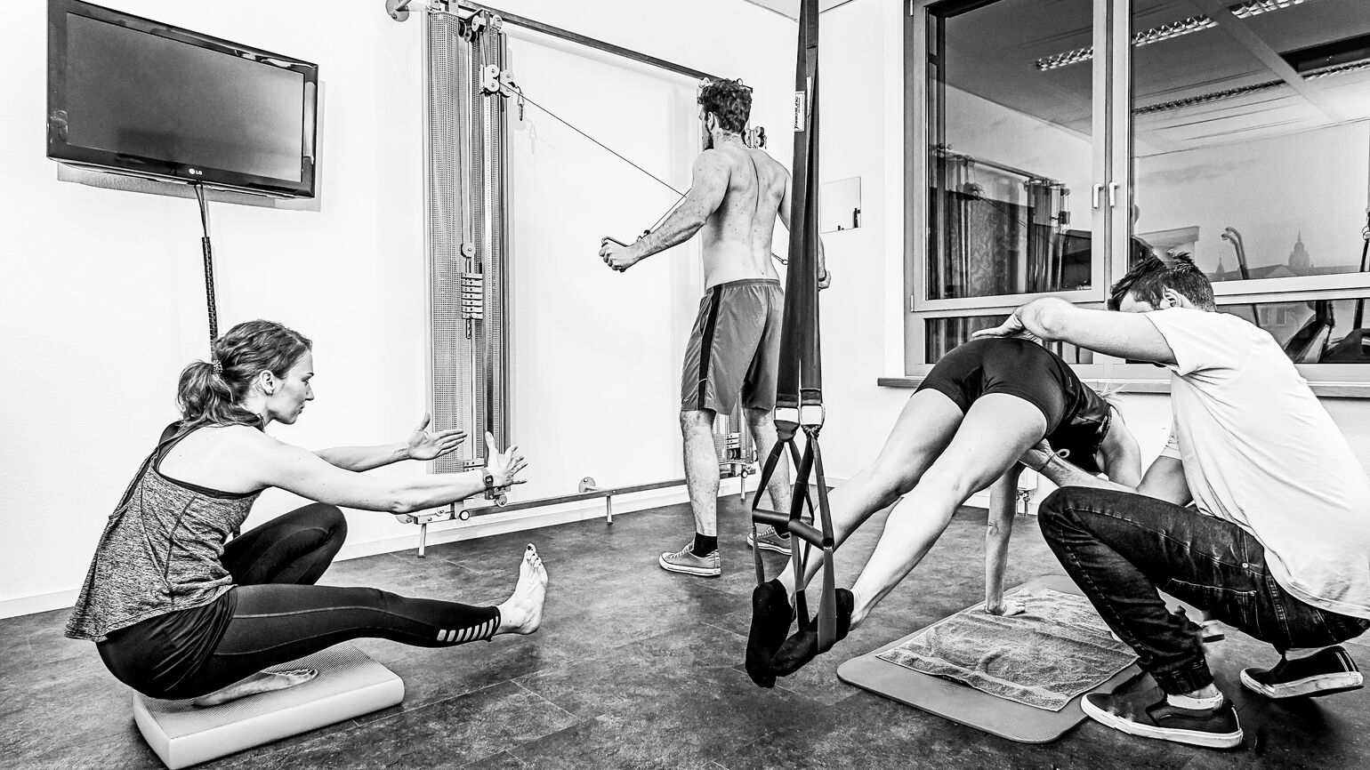 Drei Personen führen verschiedene Übungen in einem Trainingsraum durch; eine Therapeutin begleitet eine sitzende Dehnübung, ein Mann trainiert an einem Seilzug, und ein Therapeut unterstützt eine Patientin bei einer Übung in einer Schlingentrainer-Aufhäng